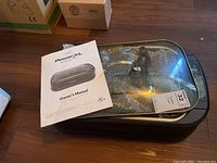 PowerXL smokeless grill with glass lid and owner's manual placed on wooden floor, grill covered partially in protective bubble wrap.