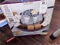 Box showing front image and branding of the stainless steel electric s'mores maker surrounded by marshmallows, chocolate and graham crackers.