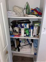 Shelving unit packed with white and patterned towels, green and white iron, colorful folded textiles, hygiene products including toothpaste and deodorants, light bulbs, and bathroom accessories.
