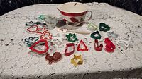Photo showing full spread of all items with mixing bowl, eggnog cup, spatula, and many colorful cookie cutters shaped as stars, hearts, stockings, trees, and more laid on a lace tablecloth.