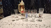 Lot showing the ceramic Tivoli advertising stein and assortment of 9 beer glasses on lace tablecloth.