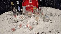 Overview of the entire vintage Coors barware collection including various glasses, mugs, and a red metal serving tray with Coors branding on a lace tablecloth.