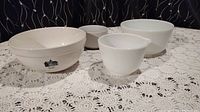 Four white mixing bowls displayed on a lace tablecloth, two identifiable as vintage Pyrex Hamilton Beach with farmhouse graphics.