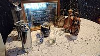Wide photo showing the Johnnie Walker Black Label mirror, silver barware items, musical decanter figurines, shot glasses, and ceramic mug arranged on a lace tablecloth.