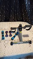 Under desk pedal exerciser and four pairs of light dumbbell weights arranged on a lace covered surface with a black patterned background.