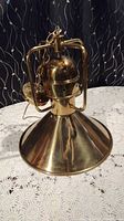 Hanging brass light fixture with a shiny conical shade, spherical central housing and metal frame support, sitting on a lace tablecloth.