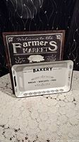 Two metal signs and a rectangular bakery tray arranged on a lace tablecloth against a dark background.