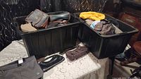 Two large black plastic totes containing various bags and purses including leather and fabric items, bags folded or placed loosely inside