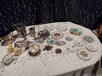 Photo showing the full assortment of silver plated and collectible plates on a lace tablecloth. Includes silver plated pitchers, trays, creamer, gravy boats, and multiple decorative plates with various scenic and floral motifs.