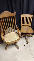 Two vintage wooden office chairs on casters, one with cane woven seat and armrests, the other solid wooden seat no armrests.