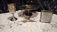 Four vintage metal items arranged on a crocheted tablecloth including a goblet, cake plate, bird figurine, and metal flask