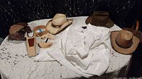 Overview of lot items on lace-covered table including three adult hats, white shirt, kids boots, and wooden accessories.