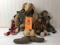 Wide group photo showing five folk art soft fabric dolls arranged against a white beadboard wall. Tags attached to four dolls.
