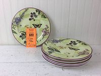 Six stacked plates with one plate standing behind showing the full floral and butterfly design with bees on a light green background and burgundy rim.