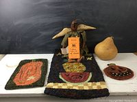 View of three wool needlework items, folk art angel with black cat, and dried gourd including rectangular fruit mat and pumpkin needlework rugs