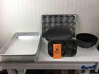 Overview of main bakeware pans including three large covered sheet pans, muffin pan, tart pan, and flower-shaped tube pan on table.