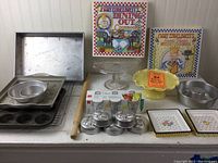 Photo showing bakeware items including metal pans, mini muffin tins, yellow and glass cake stands, two Mary Engelbreit cookbooks, a rolling pin and salt and pepper shakers shaped like little teapots.