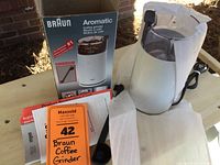 Photo of Braun Aromatic coffee grinder with clear plastic lid, coffee scoop, instructional papers, and original Braun branded box on wooden surface.
