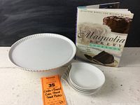 Photo of Crate & Barrel white ceramic cake stand, six small white plates, and Magnolia Bakery Cookbook on a white surface with black background.