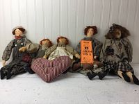 Photo showing all five folk art dolls seated side by side on white wooden floor against white paneled wall. Includes smaller Raggedy Ann and Andy dolls holding fabric heart.