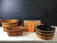 Four Longaberger baskets shown together including bread basket with bread basket brick in box, Renewal basket, small oval basket, and Petunia basket. Wooden and natural woven patterns.