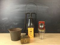 All five items shown grouped on wooden surface against a black background: Dietz kerosene lamp, Niagara Junior Guide flashlight, brass cricket box, mini pail, glass jar with handle and screw cap