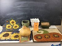 Overall view of three needlework sunflower pieces, three planters, pumpkin hooked rug, and gourd on white background with blackboard backdrop