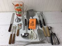 Photo shows a set of vintage kitchen gadgets arranged on a white table surface including a decorated glass pitcher with oranges, multiple spatulas, knives with wooden handles, white handled scissors, metal ladle, whisks, and peelers.