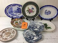Nine decorative plates arranged standing and stacked showing various designs including blue river scene, floral bouquet, blue rural scene, brown patterned plate, ivy design, and multicolor scenic plate.