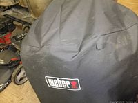 Photo of a black fabric cover with Weber logo designed for a dome-style charcoal grill.