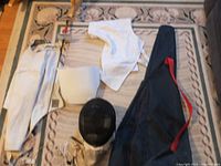 Photo of all fencing gear laid out on a carpet including jacket, pants, helmet, sword, electrical leads, protective undergarments, and carrying bag.
