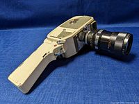 Side view of vintage Mamiya 8mm movie camera with extended pistol grip handle and attached Chinon zoom lens, resting on blue fabric.