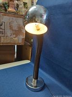 Front view of the MCM chrome flex table lamp illuminated with light on, flexible black neck curved, chrome spherical lamp head visible.