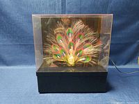 Front view of the peacock fibre optic lamp inside transparent display case showing colorful fiber optic strands arranged as peacock tail.