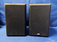 Front view of the PSB Alpha bookshelf speakers. One speaker has PSB badge on grille, the other is missing badge. Both have black fabric grilles.