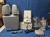 Gray Boston BA7900 subwoofer and two matching satellite speakers, three Polk Audio Monitor white surround speakers, Harman Kardon SoundSticks transparent dome speakers and control pod with cables arranged on blue background.