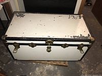 Front exterior of antique trunk showing white paint, black trim, and brass hardware.