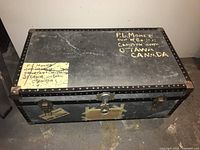Exterior view of antique black metal trunk with metal rivets, two latch locks, a central lock, and address written on top.