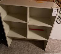 Photo showing white bookshelf with two shelves creating four compartments, placed on carpeted floor with a pink item inside one compartment.