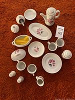 All pieces of the white toy tea set arranged on an orange fabric surface showing floral and butterfly decoration mainly in pink, blue, and yellow colors. Pieces include teapot, cups, plates, sugar bowl, creamer, salt and pepper shakers, serving dishes, and small spoons.