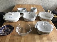 Top view of the full set showing 4 CorningWare bakeware dishes with glass lids, blue cornflower and fruit motif, plus ceramic bowls and gravy boat
