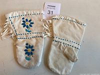 Pair of white handmade caribou skin mittens with blue beaded floral patterns and fringe, showing slight wear