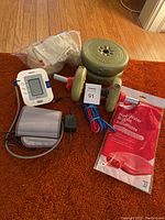 Overview photo of dumbbell with weights, Omron blood pressure monitor with cuff and adapter, resistance bands, hot water bottle in packaging, and tensor bandages on floor