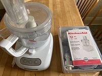 KitchenAid 12 cup food processor base with bowl, lid, feeding chute, and original manual in plastic cover on wooden table.