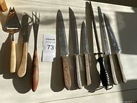 Assorted kitchen knives and tools including knives with wooden and plastic handles, a cheese cutter, and a sharpening steel laid out on a flat surface.