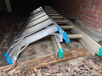 20 foot metal extension ladder lying on ground under a porch, blue end caps on rails, wooden rungs, brick wall background.