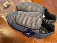 Pair of gray men's slippers for wide feet with velcro straps, shown from above on a wooden surface with packaging beneath them.
