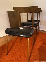 Three mid century wooden chairs with curved backrests and black vinyl seats with slight wear, legs with brass tone finish. Background shows table leaf leaning against wall.