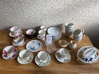 Overview of various bone china teacups, saucers, plates, and Belleek creamer and sugar on wooden surface with floral wallpaper background.