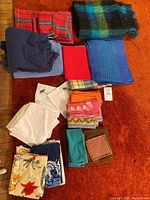 Photo showing assorted folded linens including a mohair plaid blanket, two navy sheets, and multiple colorful napkins and tablecloths folded on a carpet.